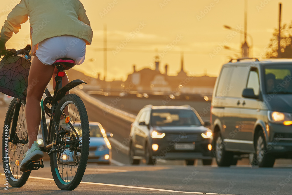 Fototapeta premium A girl in white shorts rides a bicycle next to a moving cars. A cyclist rides a bike along a cycle path at sunset next to the track.