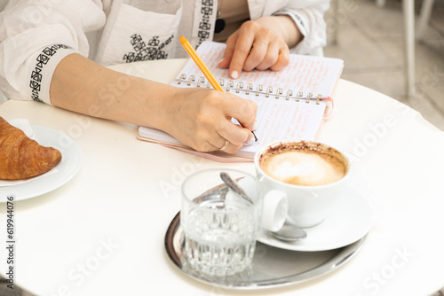 A woman writes with a pen in a notebook sitting in a cafe, a cup of coffee on the table, hands close-up