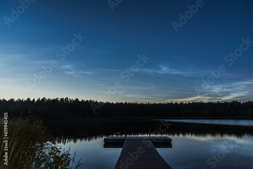 Noctilucent clouds over the forest lake in Latvia at July night. Wooden pier on foreground.