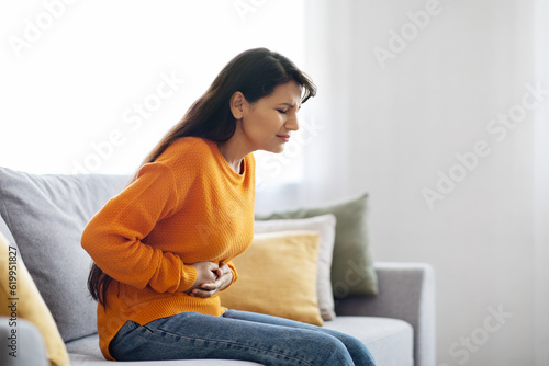 Unhappy sick young indian woman sitting on couch, touching belly photo