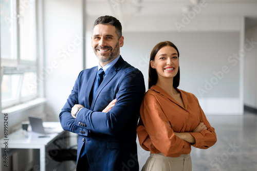 Successful businessman and businesswoman standing back to back with arms crossed and smiling at camera, office interior photo