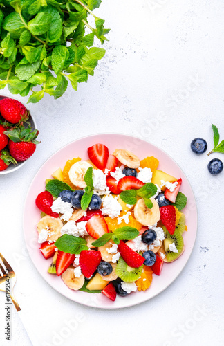 Summer fruit and berry salad with fresh strawberries, blueberries, banana, cottage cheese and mint on white table background, top view