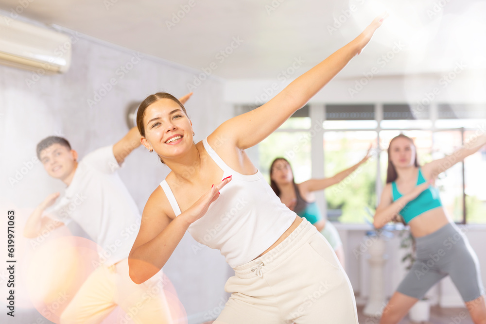 Female teenager performs movements during warm-up, limbering-up part of workout together with peers. Group of young girls and guy dance modern vogue in fitness club.