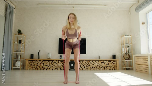 Full shot of sexy fit blonde exercising with resistance band. Home workout. Woman with slim athletic body performs bent-over row, straightening the back. High quality 4k footage