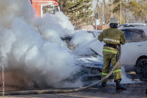 Extinguishing a burning car. A firefighter pours water from a fire hose on a burning car. Clubs of white smoke and steam. Road incident in a parking lot in the city. Dangerous job