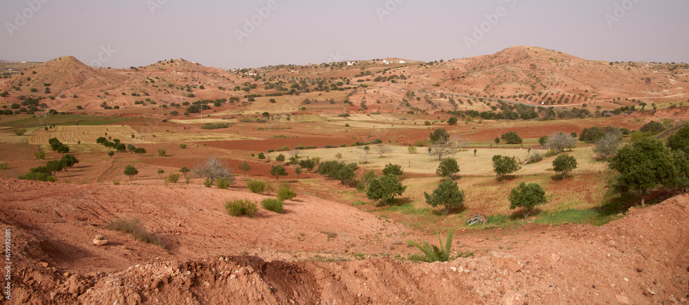 Colorful landscape of a valley with trees, bushes and hills in countryside of midland of Morocco. Spring bright sunny day. Red, orange, yellow and green colors. Vacation in Morocco. Colorful landscape of a valley with trees, bushes and hills in countryside of midland of Morocco. Spring bright sunny day. Red, orange, yellow and green colors. Vacation in Morocco.