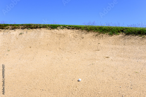 Golf ball in a sand trap, looking up at the top of the bunker and blue sky, recreation and challenge on a sunny summer day
