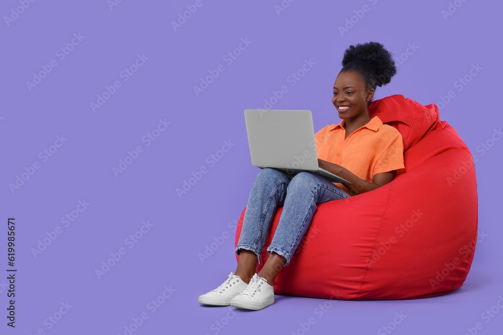 Beautiful young woman with laptop sitting on beanbag chair against purple background. Space for text Beautiful young woman with laptop sitting on beanbag chair against purple background. Space for text