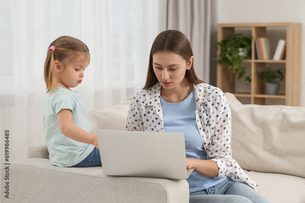 Woman working remotely at home. Little daughter bothering her mother on sofa in living room Woman working remotely at home. Little daughter bothering her mother on sofa in living room