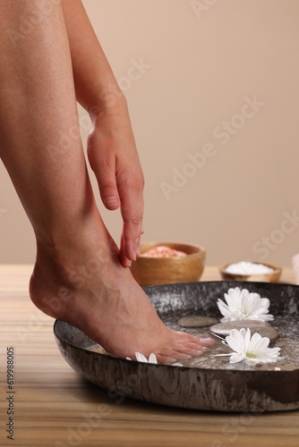 Woman soaking her foot in bowl with water, spa stones and chrysanthemum flowers on wooden surface, closeup. Pedicure procedure