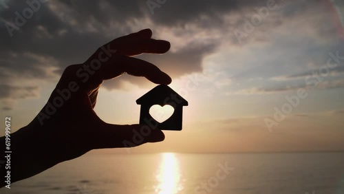 Close-up shot of woman's hand holding wooden house model on sunset and sea background. Seaside dream house concept. Choosing place for a new home. photo