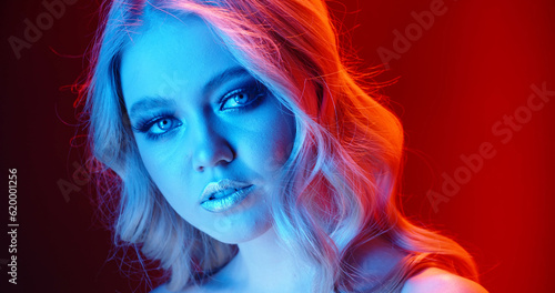 Sexy caucasian blonde girl look at camera with sensual stare, weariing bright glowing makeup, standing in red and blue neon lights - nightlife, cyberpunk concept close up portrait