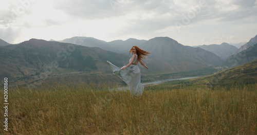Girl in light dress with red hair is walking on top of a spring mountain with scenic view. Woman in search for inspiration - freedom concept 