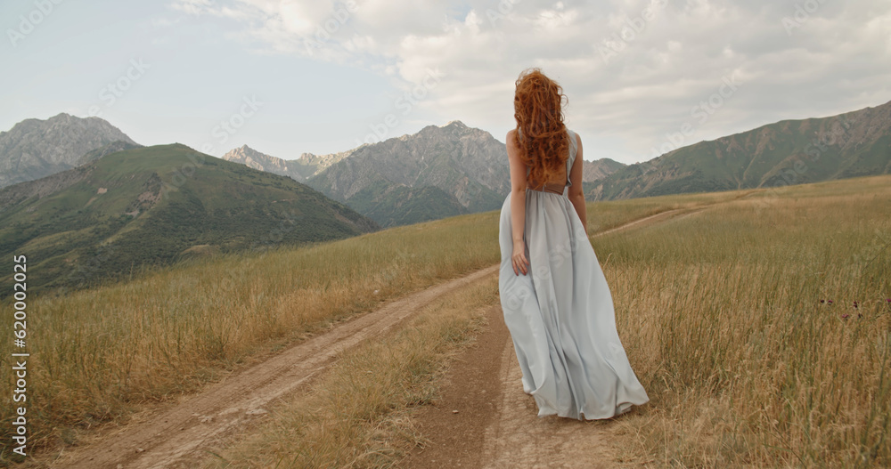 Girl with red hair walking down the wheel trails on meadow with scenic mountain background - freedom, tranquility concept .Copy space