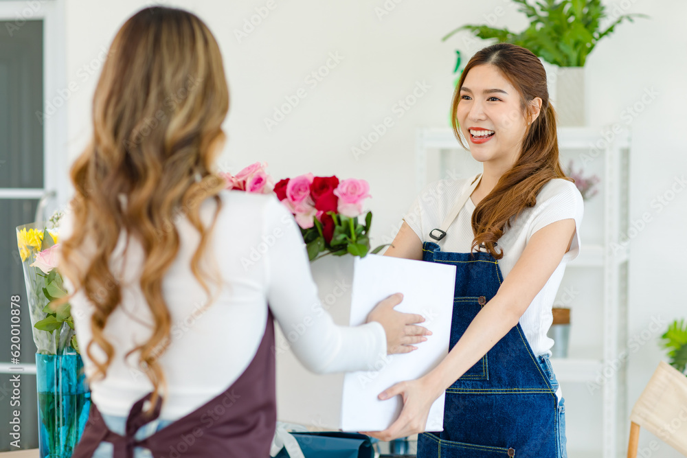 Asian happy cheerful female customer standing smiling come to flower shop buying box of red and pink roses from unrecognizable unknown florist designer owner in blurred foreground in floral studio Asian happy cheerful female customer standing smiling come to flower shop buying box of red and pink roses from unrecognizable unknown florist designer owner in blurred foreground in floral studio