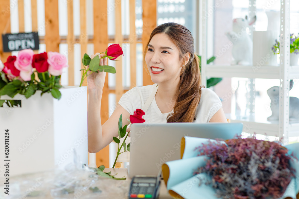 Asian happy cheerful professional female florist designer shop owner in apron sitting smiling using scissors cutting spikes decorating branch of red roses before selling in floral garden store studio Asian happy cheerful professional female florist designer shop owner in apron sitting smiling using scissors cutting spikes decorating branch of red roses before selling in floral garden store studio
