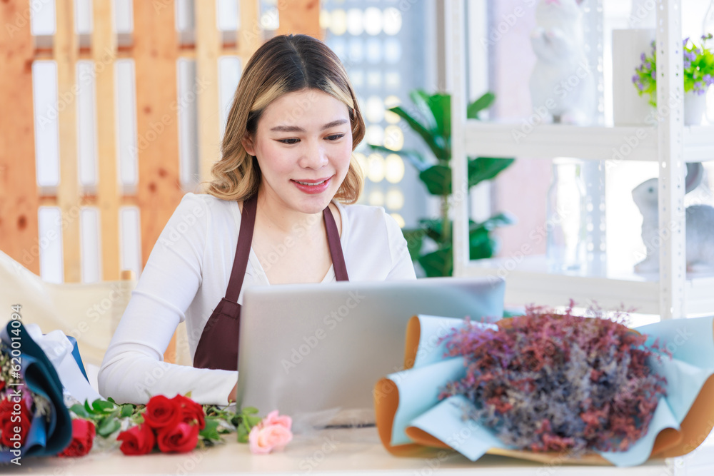 Asian happy cheerful female florist designer shop owner checking smelling quality of red roses blooming in store while using laptop notebook computer calculating finanicial target in floral studio