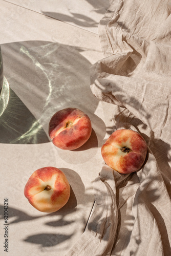 Ripe peach fruits on neutral beige tabletop, linen tablecloth, aesthetic floral sunlight shadow. Elegant sustainable lifestyle background, healthy vitamin food concept