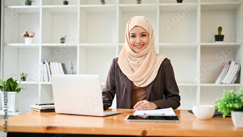 A beautiful Asian Muslim businesswoman sits at her desk in a modern private office.