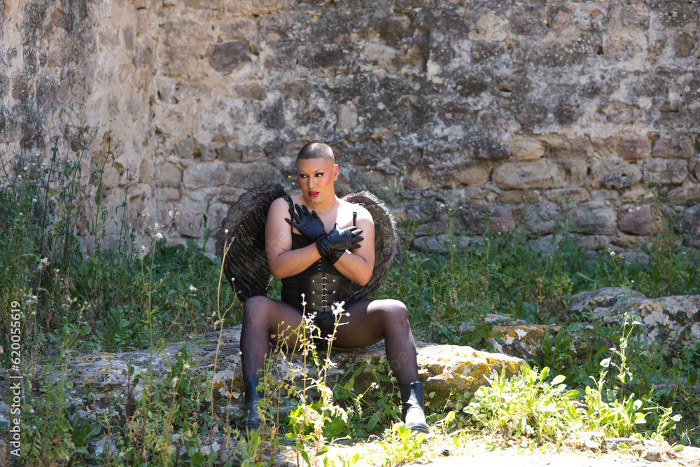 Young non-binary Latin person, wearing black make-up and black angel wings, sitting on a rock and covering his chest with his hands. Concept of diversity, homosexuality and human rights.