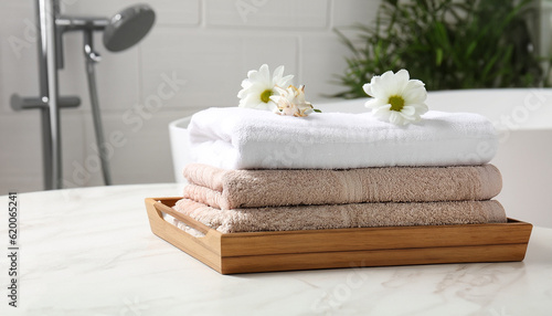 Wooden tray with stacked bath towels and beautiful flowers on white table in bathroom. Space for text