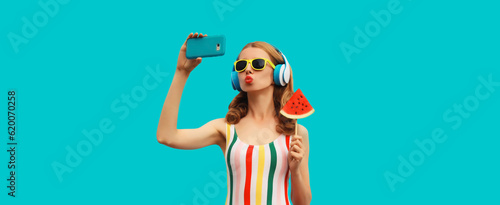 Summer portrait of cheerful happy young woman taking selfie with smartphone in headphones listening to music with juicy lollipop or ice cream shaped slice of watermelon on blue background