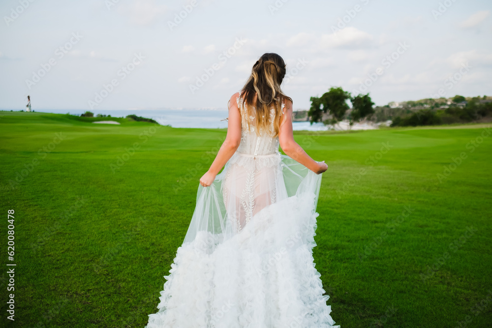 A beautiful, young girl with blond hair runs across the lawn in the light of the setting sun, in a white, stylish wedding dress, rear view.