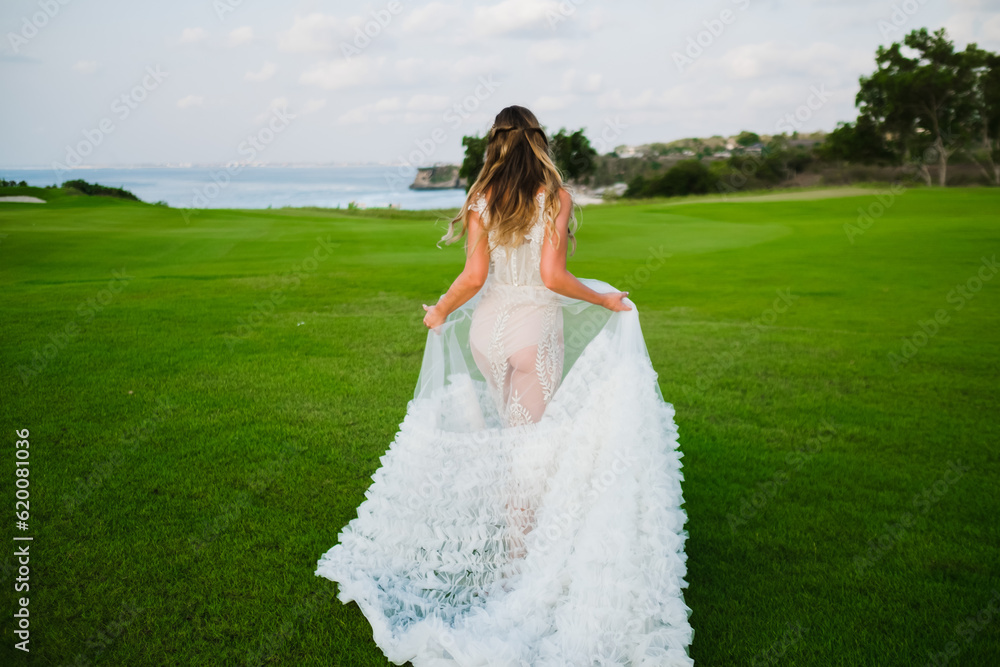 A beautiful, young girl with blond hair runs across the lawn in the light of the setting sun, in a white, stylish wedding dress, rear view. A beautiful, young girl with blond hair runs across the lawn in the light of the setting sun, in a white, stylish wedding dress, rear view.