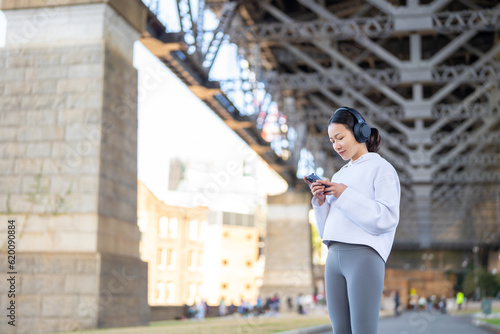 Asian woman in sportswear listening to music from headphones with mobile phone app during jogging exercise at city street in the morning. Healthy girl do sport training running workout in the city.