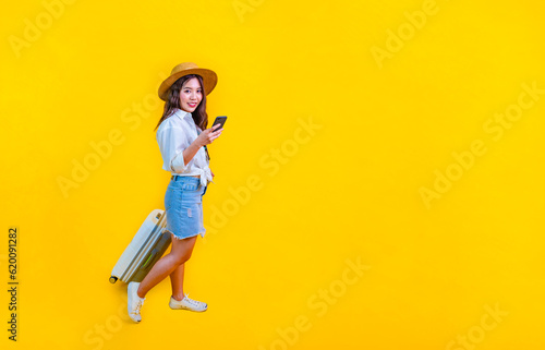 Pretty Asian woman passenger in trendy fashion is using mobile phone while carrying her luggage bag in happiness for travel and summer vacation isolated on yellow background © Akarawut