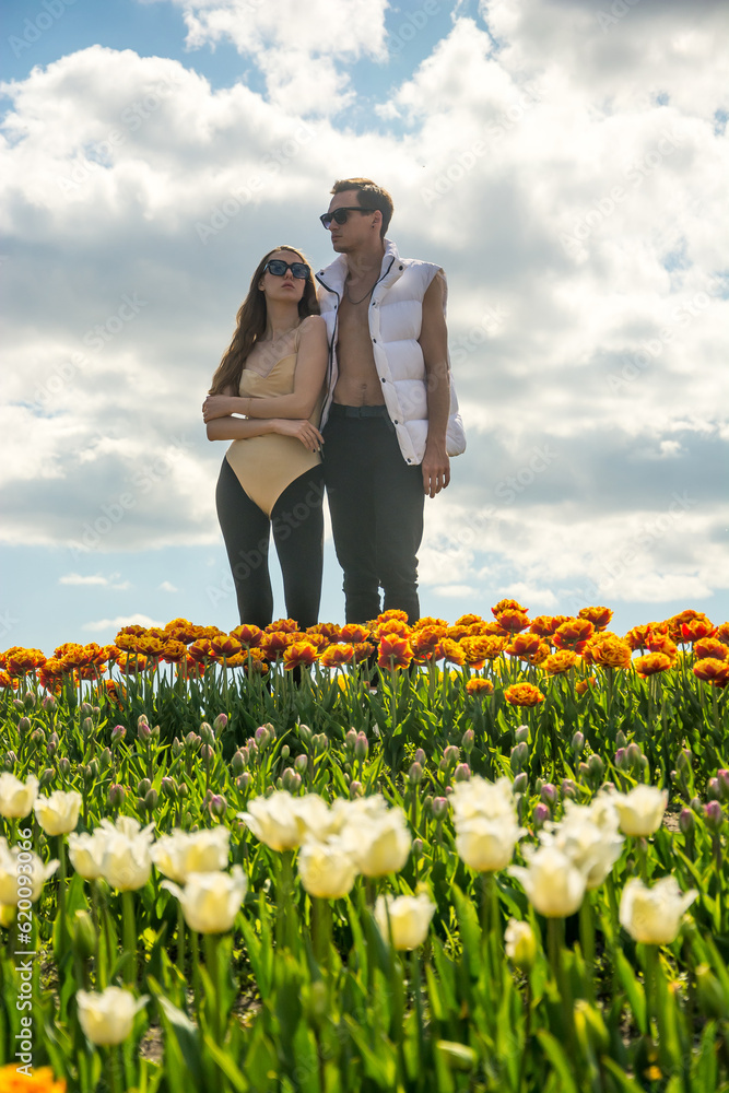 A beautiful young couple, fashion models stands in sportswear at the background of a field of colorful tulips.