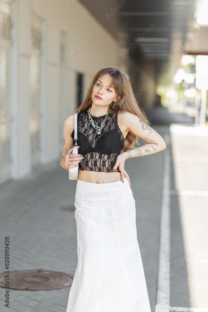 Fashion beautiful young girl in a stylish black lace top with a bra and a white skirt with a handbag walks on the street. Hipster woman with tattoos on her arms