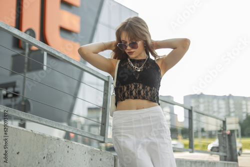 Fashionable young beautiful hipster woman model hipster with sunglasses in black fashion lace top and bra in white skirt with bag stands and poses in the city © alones