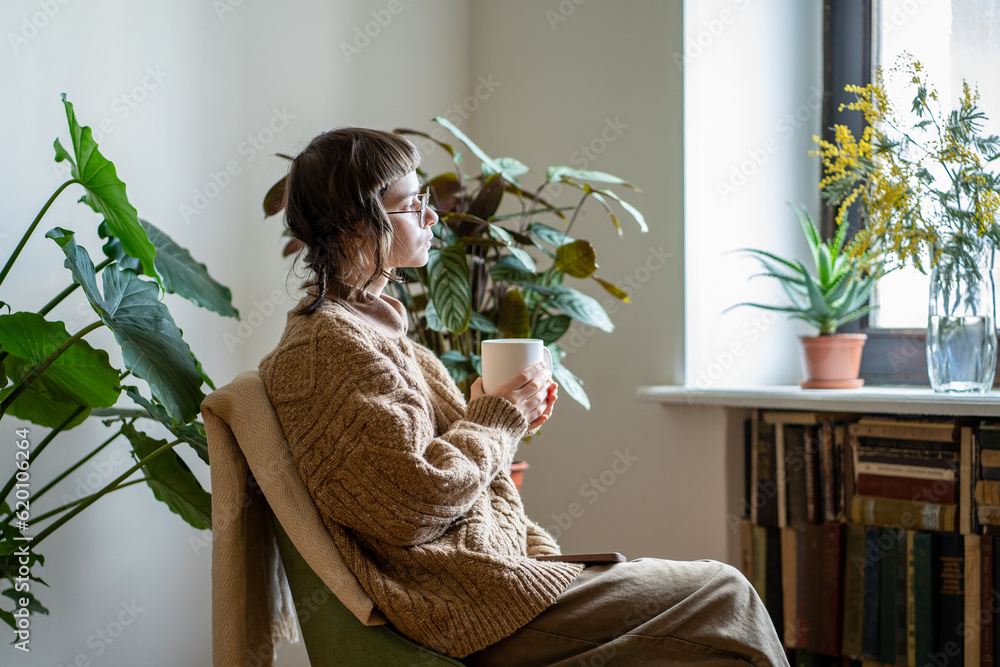 Φωτογραφία Thoughtful dreaming creative girl sits on armchair with cup coffee look out window enjoy day time in cozy place house with green plants in pots