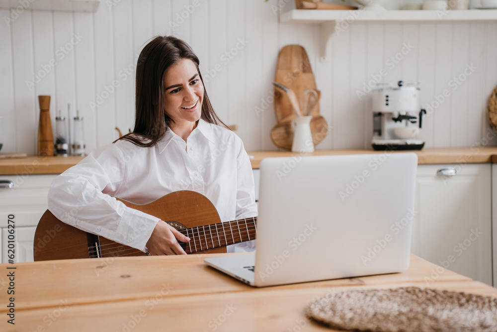 Cheerful Brazilian girl in white shirt sitting at desk with laptop holds guitar talks to teacher of guitar playing. Remote lesson, hobby. Pretty caucasian woman teaching via internet. Distant lesson Cheerful Brazilian girl in white shirt sitting at desk with laptop holds guitar talks to teacher of guitar playing. Remote lesson, hobby. Pretty caucasian woman teaching via internet. Distant lesson