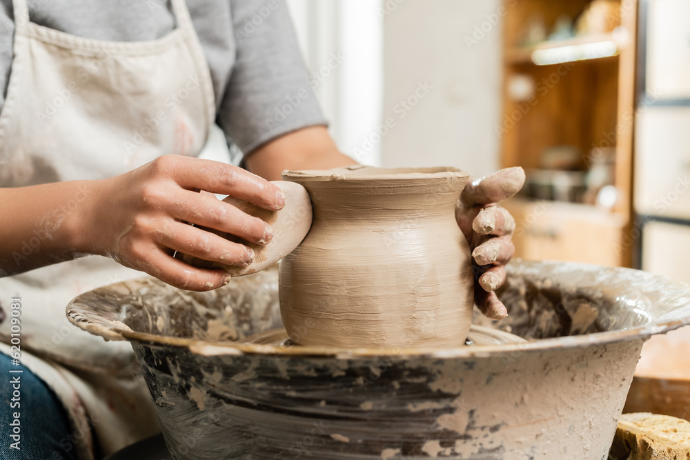 Cropped view of blurred young craftswoman in apron holding wooden scraper while making clay vase on pottery wheel in art studio at background, clay shaping and forming process Cropped view of blurred young craftswoman in apron holding wooden scraper while making clay vase on pottery wheel in art studio at background, clay shaping and forming process