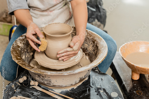 Cropped view of female potter in apron making shape of clay vase with vet sponge near wooden tools and spinning pottery wheel in ceramic workshop, clay shaping and forming process