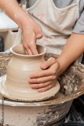 Cropped view of young female artisan in apron making shape of clay vase and working with spinning pottery wheel on table in ceramic workshop, pottery creation process