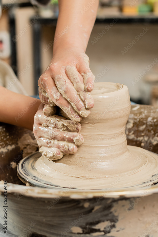 Cropped view of young female ceramicist molding wet clay on spinning pottery wheel while working in blurred ceramic workshop at background, skilled pottery making concept