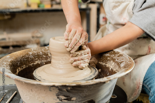 Cropped view of blurred female ceramicist in apron molding wet clay and working on spinning pottery wheel in art ceramic studio, skilled pottery making concept