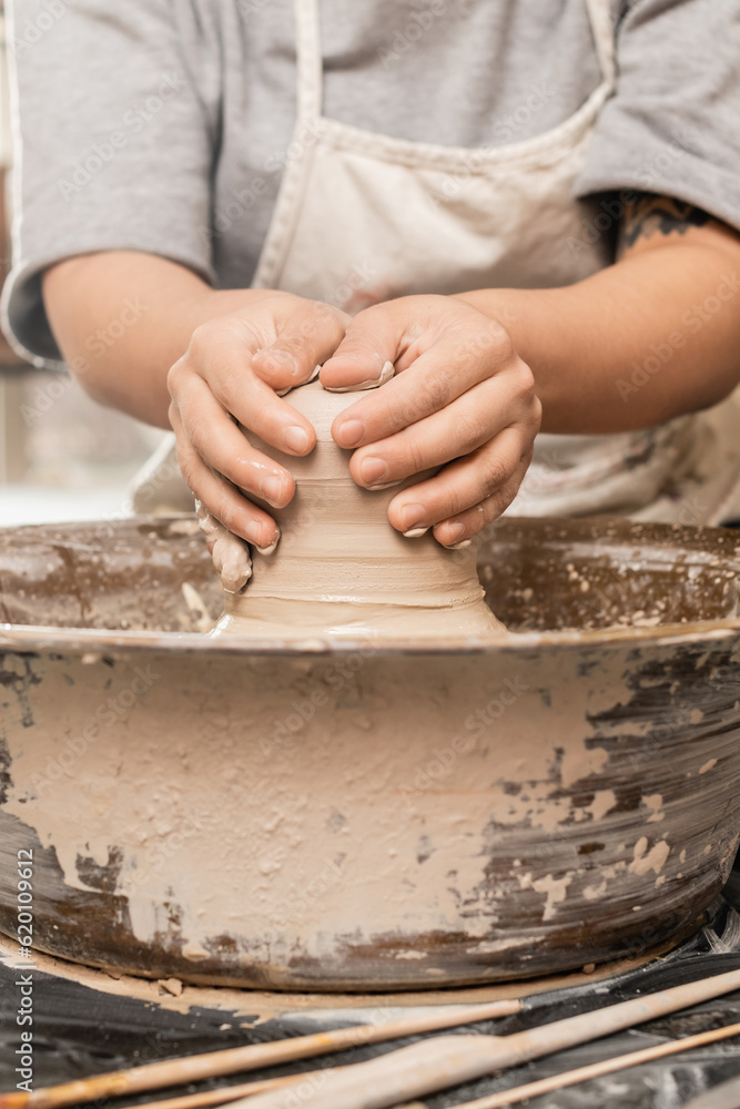Cropped view of blurred and tattooed craftswoman in apron molding wet clay while working on pottery wheel near tools on table in ceramic studio, clay sculpting process concept Cropped view of blurred and tattooed craftswoman in apron molding wet clay while working on pottery wheel near tools on table in ceramic studio, clay sculpting process concept