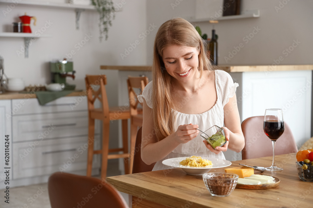 Young woman adding pesto to tasty pasta in kitchen