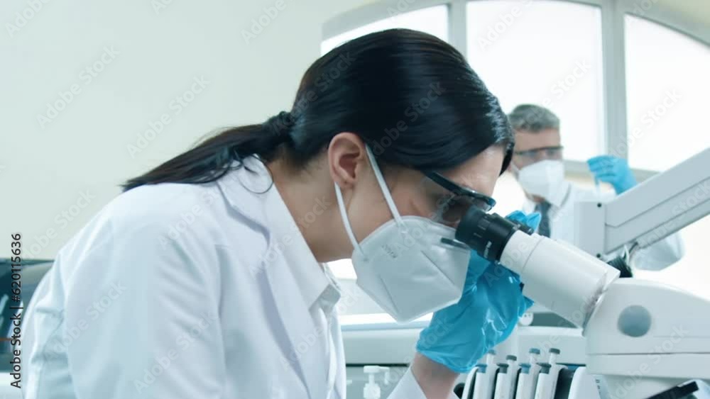 Shoot from side. Young female scientist in lab coat, safety glasses and medical mask. Woman conducts research in laboratory. Concentrated woman looking into microscope on workflow background. Shoot from side. Young female scientist in lab coat, safety glasses and medical mask. Woman conducts research in laboratory. Concentrated woman looking into microscope on workflow background.
