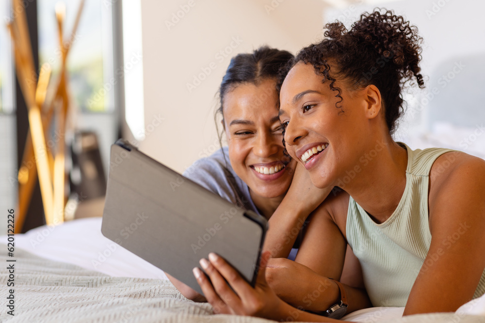Happy biracial lesbian couple using tablet on bed in bedroom