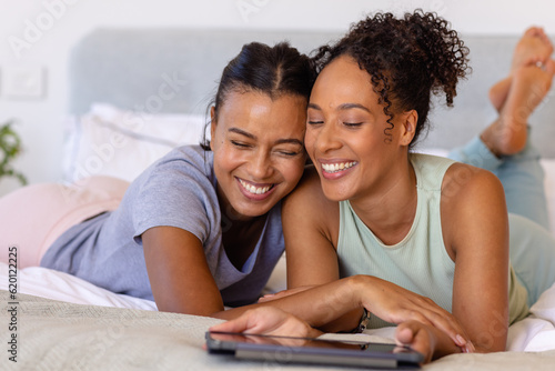 Happy biracial lesbian couple embracing and using tablet on bed in bedroom