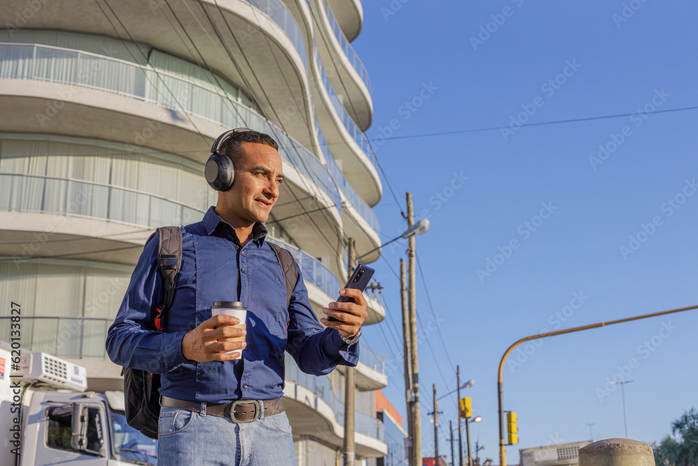 Young latin man with headphones looking at his mobile phone and holding a paper cup with coffee in his hand with copy space. Young latin man with headphones looking at his mobile phone and holding a paper cup with coffee in his hand with copy space.