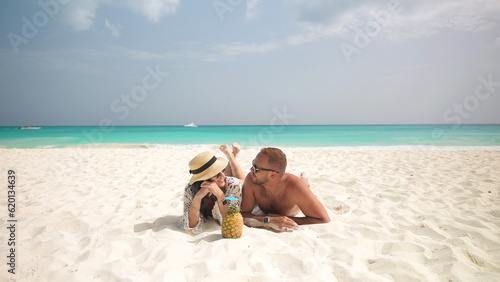 Latin couple lies and smiling on the white sand on the tropical beach. Pineapple cocktail and paradise pleasure against the background of the turquoise sea. photo