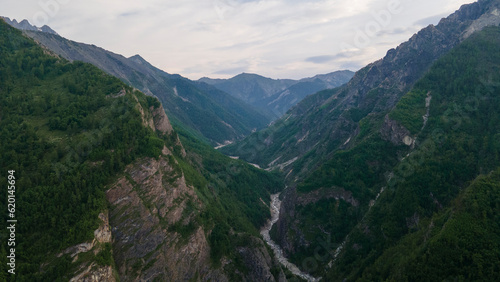 Aerial view of the high waterfall and fast mountain river in valley of the rocky mountain ranges, on which snow lies in places at summer cloud sunrise, color