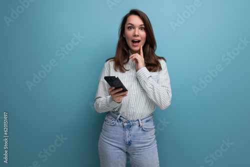 surprised european young brunette female adult in a striped shirt and jeans with a smartphone in her hands on a studio background