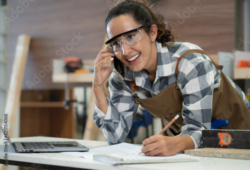 Beautiful Latin woman carpenter using mobile phone talking to customer for woodworking ordering in carpenter's shop. Female hispanic woodworker writing order note. Feminism in carpentry industry.